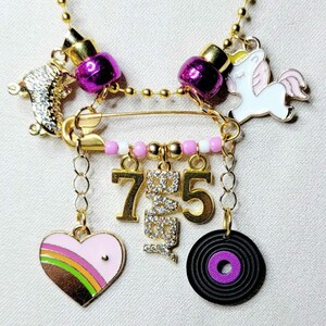 May include: Gold charm necklace with a safety pin, pink and white beads, a unicorn charm, a roller skate charm, a heart charm with a rainbow stripe, a record charm, and two gold charms with rhinestones that say "75".