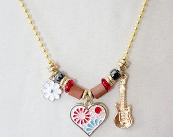 May include: A gold-colored necklace with a ball chain. The necklace features a white flower charm, a heart-shaped pendant with a floral design, and a guitar charm. The heart pendant has red, pink, and blue accents.