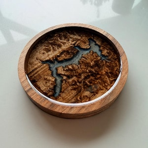 Puede incluir: Posavasos redondo de madera con un patrón de vetas de madera natural y una incrustación de resina azul. El posavasos tiene un borde plateado y está diseñado para proteger las superficies de la condensación.