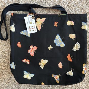 K&ouml;nnte beinhalten: Schwarze Tragetasche mit schwarzem Trageriemen und einem wei&szlig;en Etikett mit der Aufschrift "Little Ladybug" und einem Schmetterlingsdesign. Die Tasche ist mit einem Muster aus bunten Schmetterlingen bedeckt.