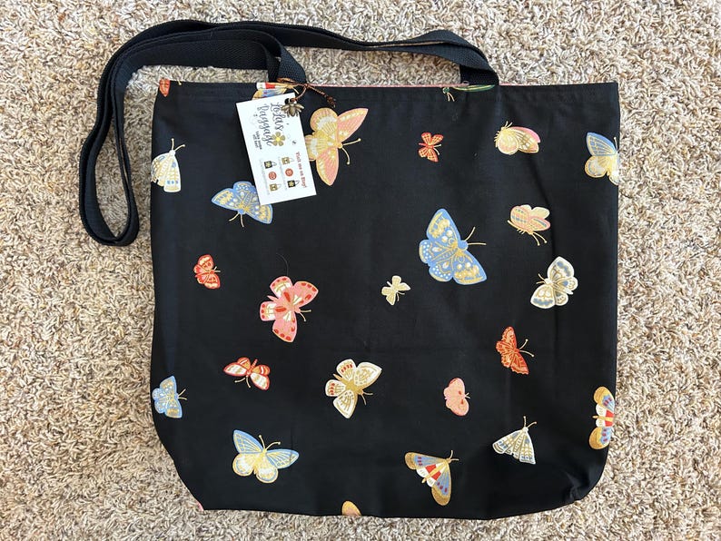 K&ouml;nnte beinhalten: Schwarze Tragetasche mit schwarzem Trageriemen und einem wei&szlig;en Etikett mit der Aufschrift "Little Ladybug" und einem Schmetterlingsdesign. Die Tasche ist mit einem Muster aus bunten Schmetterlingen bedeckt.