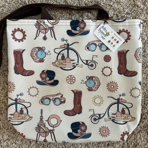 May include: Cream-colored tote bag with a brown strap, featuring a repeating pattern of steampunk-themed illustrations in brown, blue, and tan. The design includes gears, boots, hats, and bicycles. A tag with text is attached.