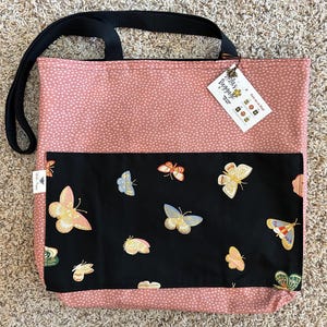 K&ouml;nnte beinhalten: Eine rosa Tragetasche mit schwarzen Punkten und einer schwarzen Tasche mit einem Schmetterlingsmuster. Die Tasche hat einen schwarzen Griff und ein Etikett mit der Aufschrift "Bella's Baggage".