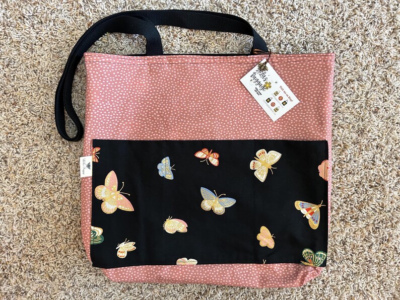 K&ouml;nnte beinhalten: Eine rosa Tragetasche mit schwarzen Punkten und einer schwarzen Tasche mit einem Schmetterlingsmuster. Die Tasche hat einen schwarzen Griff und ein Etikett mit der Aufschrift "Bella's Baggage".