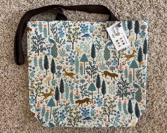 May include: A cream-colored tote bag with a repeating pattern of animals and foliage in blue, green, and orange. The design includes deer, leopards, peacocks, and trees. The bag has a brown strap and a tag.