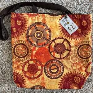 May include: A tote bag with a brown strap and a repeating gear pattern in shades of orange, yellow, and brown. The bag has a tag attached with the text "Lola's Baggage".