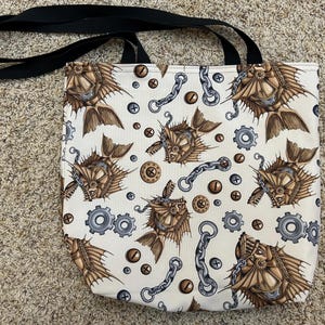 May include: A white canvas tote bag with a black strap. The bag features a repeating pattern of brown and gold fish with gears and chains.