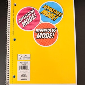 May include: A yellow spiral notebook with three circular stickers that read "HYPERFOCUS MODE!" in white text. The stickers are in red, blue, and orange. The notebook has a perforated edge and is labeled "PEN+GEAR".