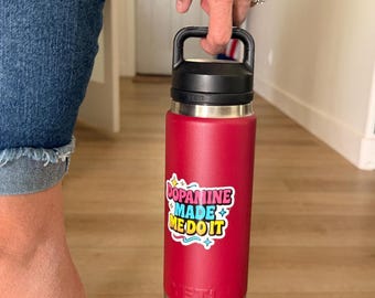 May include: A red insulated water bottle with a black handle and lid. A colorful sticker on the bottle reads "Dopamine Made Me Do It." The bottle is made of stainless steel and has a silver base.
