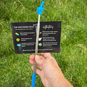 May include: A blue and silver "Shoosher Stick" with a pointing finger design. The stick is held in front of a card with instructions and the text "The Shoosher Stick". The background is green grass.