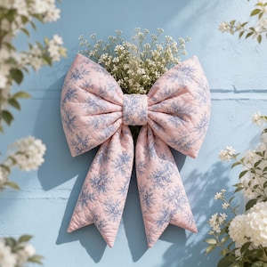 May include: A large, quilted fabric bow featuring a soft pink background with a repeating blue floral pattern. The bow is constructed with two wide loops and two long, pointed tails, and it holds a small bouquet of white baby's breath flowers.
