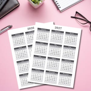 May include: Two white 2026 and 2027 yearly calendars with monthly grids, displayed on a pink surface. The calendars are surrounded by office supplies, including a pen, glasses, and a small plant.