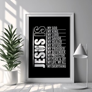 Puede incluir: Impresión en blanco y negro enmarcada con el texto "JESUS IS" y una lista de atributos que incluyen "MY GOD", "MY KING" y "MY SAVIOR". La impresión está en un marco blanco, exhibida sobre una superficie blanca, con una planta en maceta y una lámpara en el fondo.
