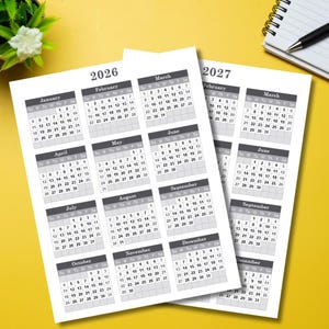 May include: Two white paper calendars for the years 2026 and 2027, with monthly grids in a simple, readable format. The calendars are on a yellow surface, with a small plant and a notepad in the background.