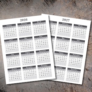 May include: Two printed calendar sheets for the years 2026 and 2027. Each sheet displays a monthly calendar grid with dates, days of the week, and month names in a simple, readable font. The background is a light wood grain.