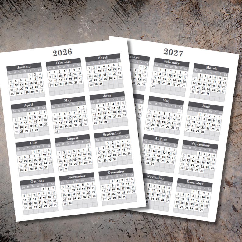 May include: Two printed calendar sheets for the years 2026 and 2027. Each sheet displays a monthly calendar grid with dates, days of the week, and month names in a simple, readable font. The background is a light wood grain.
