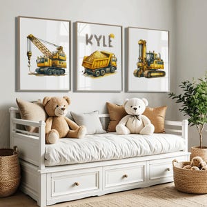 Könnte beinhalten: Drei gerahmte Aquarelldrucke mit Baumaschinen: ein Kran, ein Muldenkipper mit dem Namen "KYLE" und ein Bagger. Die Drucke befinden sich in einem Kinderzimmer, über einem weißen Tagesbett mit Teddybären und Kissen.