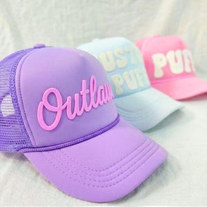 May include: Three brightly coloured trucker caps. The first is lavender with the word "Outlaw" in pink script. The second is light blue with "Puff" in white. The third is pink with "Puff" in white. All have mesh backs.