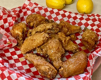 May include: A basket of fried chicken wings on red and white checkered paper. Three yellow lemons are in the background. The wings are golden brown and appear crispy.