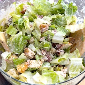 May include: A Caesar salad in a clear glass bowl. The salad contains crisp, green romaine lettuce, croutons, bacon bits, and creamy dressing. A wooden spoon is visible. A healthy and delicious meal.