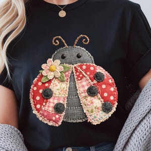 May include: Black t-shirt featuring a whimsical ladybug design. The ladybug has red polka-dot wings, black spots, and a floral and patchwork pattern. A fabric flower is on the ladybug's head. The shirt is a casual, everyday wear item.
