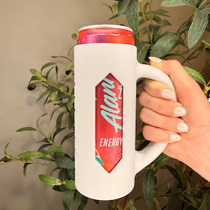 May include: A white, mug-shaped can cooler holds a red and teal energy drink can. The cooler has a handle and a cutout revealing the drink's label, which reads "Alani Energy". The background features green foliage.