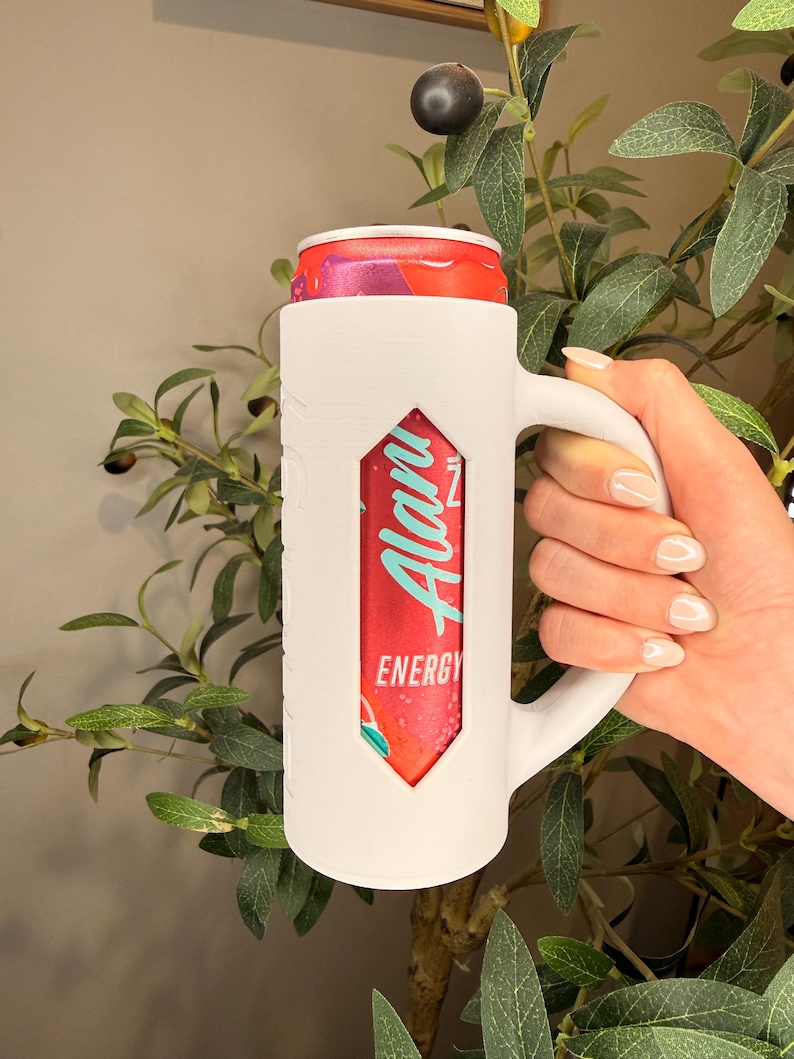 Puede incluir: Un enfriador de latas blanco en forma de taza sostiene una lata de bebida energ&eacute;tica roja y turquesa. El enfriador tiene un asa y un recorte que revela la etiqueta de la bebida, que dice "Alani Energy". El fondo presenta follaje verde.