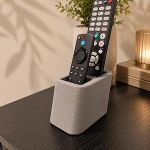 May include: A light gray, textured, rectangular remote control holder with rounded corners, containing two black remote controls of different sizes. The holder has two distinct slots to keep the remotes upright and organized for easy access.