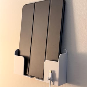 Puede incluir: Un soporte de tableta blanco montado en la pared con una tableta gris y un cable de carga blanco. La tableta está en una funda gris. El soporte tiene dos compartimentos y está montado en una pared blanca.