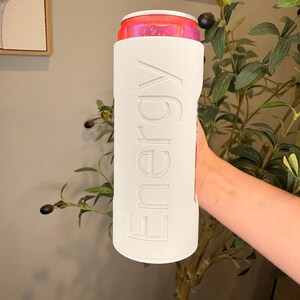 Puede incluir: Un enfriador de latas blanco con la palabra "Energy" en relieve en el lateral, que sostiene una lata rosa y roja. El enfriador es cil&iacute;ndrico y parece estar hecho de un material r&iacute;gido. La lata es parcialmente visible en la parte superior.