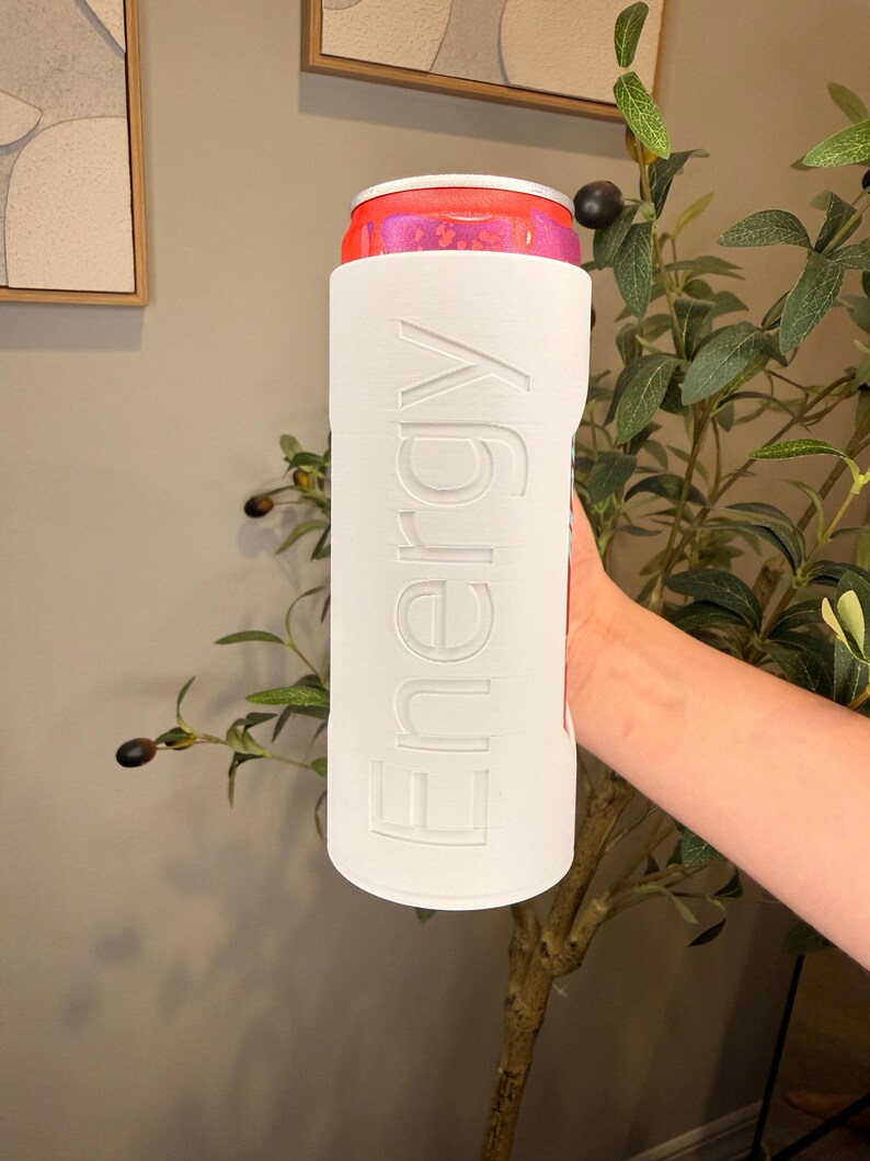 Puede incluir: Un enfriador de latas blanco con la palabra "Energy" en relieve en el lateral, que sostiene una lata rosa y roja. El enfriador es cil&iacute;ndrico y parece estar hecho de un material r&iacute;gido. La lata es parcialmente visible en la parte superior.