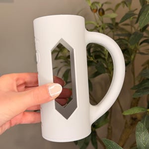 Puede incluir: Una taza de cer&aacute;mica blanca con un dise&ntilde;o de corte geom&eacute;trico &uacute;nico. La taza tiene un asa grande y una forma cil&iacute;ndrica. La taza est&aacute; sostenida por una persona. La taza es de un color claro y neutro.
