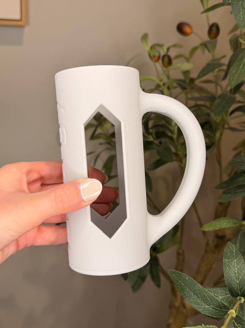 Puede incluir: Una taza de cer&aacute;mica blanca con un dise&ntilde;o de corte geom&eacute;trico &uacute;nico. La taza tiene un asa grande y una forma cil&iacute;ndrica. La taza est&aacute; sostenida por una persona. La taza es de un color claro y neutro.