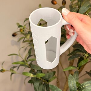 Puede incluir: Taza blanca con un dise&ntilde;o de corte geom&eacute;trico. La taza tiene un asa y una forma cil&iacute;ndrica. La taza est&aacute; sostenida por una mano. La taza tiene una superficie texturizada y est&aacute; rodeada de hojas verdes.