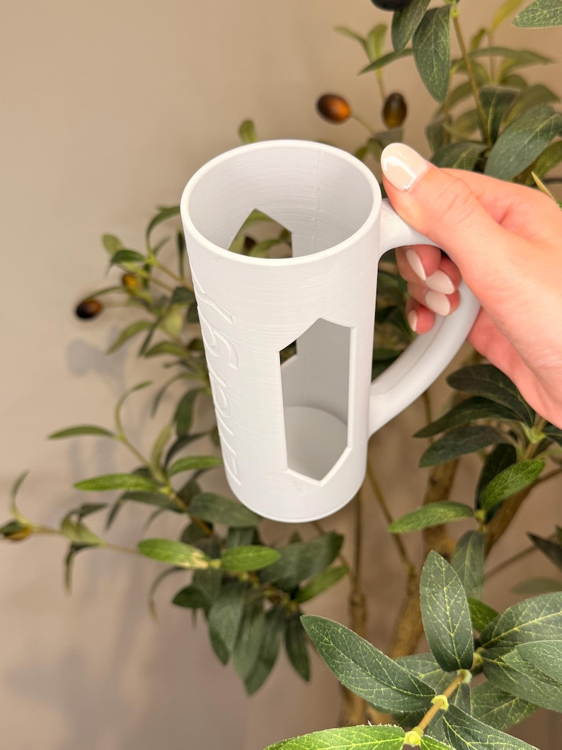 Puede incluir: Taza blanca con un dise&ntilde;o de corte geom&eacute;trico. La taza tiene un asa y una forma cil&iacute;ndrica. La taza est&aacute; sostenida por una mano. La taza tiene una superficie texturizada y est&aacute; rodeada de hojas verdes.