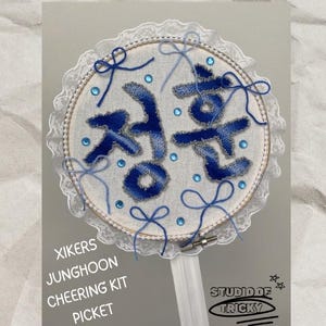 Puede incluir: Un aro bordado azul y blanco con la palabra coreana "Cheer" en hilo azul. El aro está decorado con encaje blanco y lazos azules. El texto "Xikers Junghoon Cheering Kit Picket" está escrito debajo del aro. El texto "Studio of Tricky" está escrito en un círculo negro con estrellas debajo del aro.