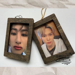 May include: Two wooden framed photos with images of people. The frames are rectangular with a dark brown finish. One frame has a silver chain, and the other has a gold chain. The text "STUDIO OF TRICKY" is visible on one frame.