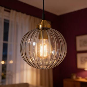 May include: A clear, ribbed glass pendant light with a brass-colored top and a vintage-style Edison bulb. The light hangs from a black cord, creating a warm, inviting glow. The spherical shape adds a touch of elegance to any room.