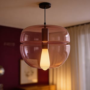 May include: A modern, spherical pendant light fixture with a transparent, dusty rose-colored glass shade. The light has a cylindrical, dark brown metal stem and a warm, white LED bulb. The fixture is suspended from a black ceiling mount.