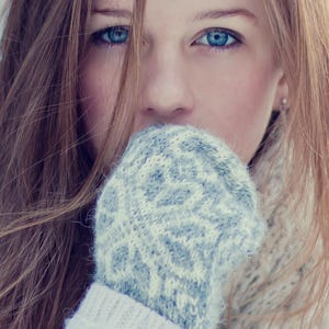 May include: A person with long brown hair and striking blue eyes, wearing a light gray knitted mitten with a geometric pattern. A matching sweater and scarf are also visible, creating a cozy winter look.