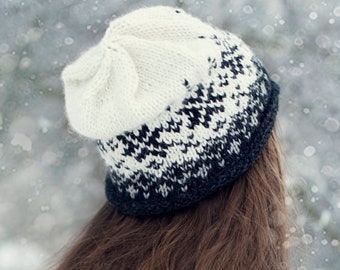 Könnte beinhalten: Eine Frau mit langen braunen Haaren trägt eine weiße und schwarze Strickmütze und einen grauen Pullover. Der Hintergrund ist eine verschneite Winterlandschaft.
