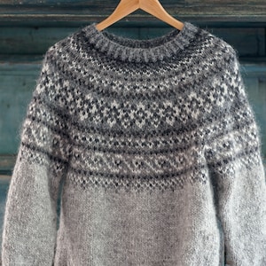 以下が含まれることがあります： 木製のハンガーに掛けられた、丸いネックラインのグレーの長袖セーター。セーターは、白、黒、グレーの幾何学模様のパターン化されたヨークが特徴です。袖と下半身は無地のグレーです。