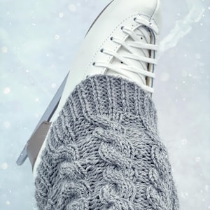 May include: A close-up of a white ice skate with a grey knitted cable-knit sock. The skate is on a white surface with a light blue tint.