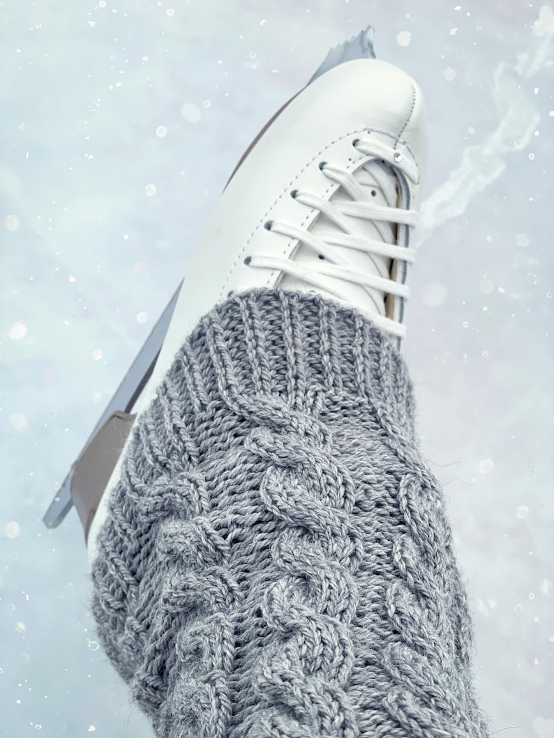 May include: A close-up of a white ice skate with a gray knitted cable-knit sock. The skate is on a white surface with a light blue tint.