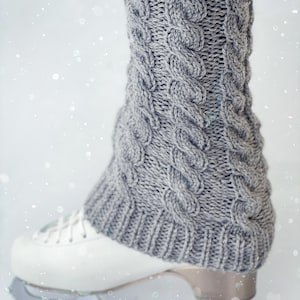 May include: Gray knitted leg warmers with a cable knit pattern, worn over white ice skates.
