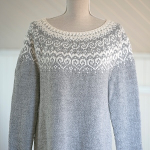 Könnte beinhalten: Ein grauer und weißer Strickpullover mit einem geometrischen Muster um den Halsausschnitt. Der Pullover hat lange Ärmel und eine lockere Passform.