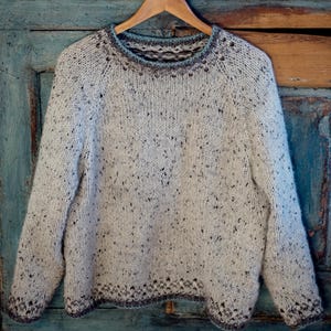 Könnte beinhalten: Ein weißer Pullover mit einem gesprenkelten Muster und einem blauen und braunen Saum an Hals und Saum.