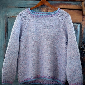 Può includere: Un maglione di maglia viola chiaro con maniche lunghe, appeso a una gruccia di legno. Il maglione ha uno scollo tondo ed è rifinito con accenti blu e rosa sullo scollo e sull'orlo. Il testo "KATRINE H." è visibile in basso.