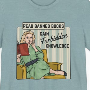 May include: A light blue t-shirt with a vintage-style illustration of a woman reading a book. The text on the shirt reads "Read Banned Books Gain Forbidden Knowledge."