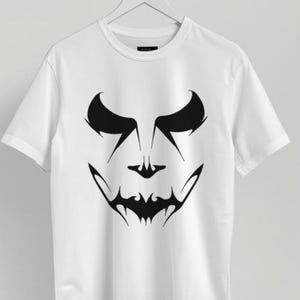 Könnte beinhalten: Weißes T-Shirt mit einer schwarzen Grafik eines stilisierten Gesichts. Das Design weist scharfe, eckige Elemente auf, darunter kräftige Augenbrauen, spitze Augen und ein gezackter Mund, wodurch eine gruselige, Halloween-Thematik entsteht.